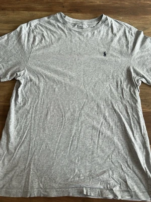 Grå t-shirt från Polo Ralph Lauren - Barnstrolek i XL. Klassisk grå t-shirt från Polo Ralph Lauren med rund halsringning och den ikoniska broderade loggan på bröstet. Tillverkad i mjuk bomull och har en normal passform. Perfekt basplagg för alla tillfällen.