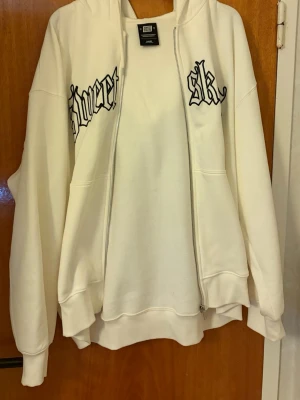 Vit hoodie med tryck och dragkedja - Vit hoodie med dragkedja och huva. På bröstet finns svart tryck i gotisk stil med texten 'Sweet' och 'sk'. Hoodien har två fickor framtill och ribbade muddar vid ärmar och nederkant. Tillverkad i mjukt sweatshirtmaterial, perfekt för en avslappnad streetwear-look.