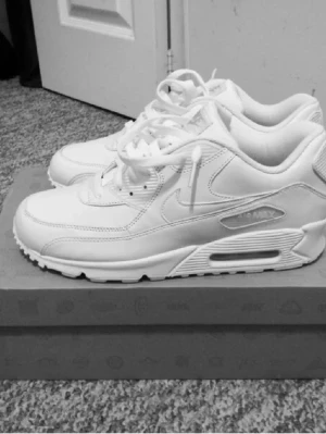 Helvita Nike Air Max 90 sneakers - Säljer ett par helvita Nike Air Max 90 sneakers med klassisk chunky sula och synlig Air-enhet. Skorna har en stilren design med läder- och meshdetaljer samt Nike-logga på sidan. Perfekta för dig som gillar en clean och sportig look.