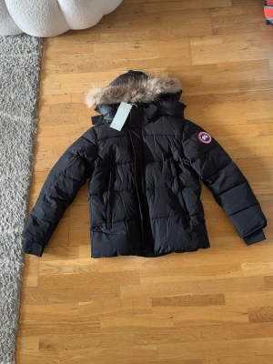 Svart vinterjacka från Canada goose - Snygg svart vinterjacka med huva och pälskant från Canada goose. Jackan har ett klassiskt quiltat mönster och en patch på ärmen. Perfekt för kalla vinterdagar och håller dig varm med sin tjocka vaddering. Dragkedja framtill och praktiska fickor, det står storlek S men passar både M och S då den är lite större än storlek S.