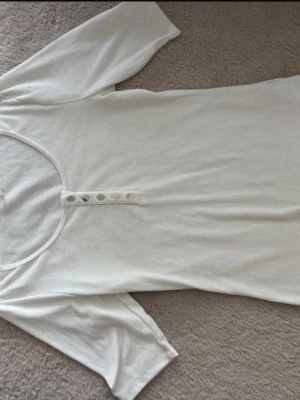 Vit topp med knappar fram - En enkel vit topp med rund halsringning och dekorativa knappar framtill. Modellen har korta ärmar och är gjord i mjukt bomullsmaterial. Perfekt att styla med jeans eller kjol för en clean look.