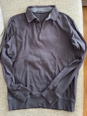 Stålgrå/blå långärmad pikétröja Tommy Hilfiger - Snygg långärmad pikétröja från Tommy Hilfiger med klassisk krage och knappslå. Tröjan har diskret logga broderad på bröstet och ribbade muddar vid ärmslut och nederkant. Perfekt för en avslappnad men stilren look.