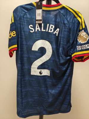 Arsenal bortatröja 26/27 spelar version Saliba #2 - Mörkblå Arsenal bortatröja med gula och röda detaljer, spelarnamn Saliba och nummer 2 på ryggen. Premier League-märke och 'No room for racism' på ärmen. Adidas-logga och Emirates Fly Better på bröstet. Tillverkad i lätt funktionsmaterial.