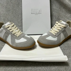 Maison Margiela gråvita sneakers - Maison Margiela sneakers i vitt och ljusgrått med ovandel i läder och mocka. Klassisk låg modell med beige snörning och gummisula i brunt. Snygg och stilren design som passar till det mesta. Diskret logga på plösen.