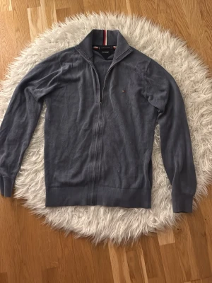 Blå ziptröja från Tommy Hilfiger - Mjuk blå ziptröja från Tommy Hilfiger i pima bomullsblandning med cashmere. Tröjan har hög krage, dragkedja framtill och diskret logga på bröstet. Perfekt för en stilren och avslappnad look.