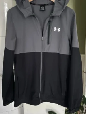 Svart och grå vindjacka Under Armour - Snygg vindjacka från Under Armour i svart och grått med huva och hel dragkedja. Jackan har en bröstficka med dragkedja, elastiska muddar och logga på bröstet. Perfekt för blåsiga dagar och sportiga outfits.