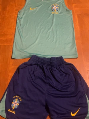 Brasilien träningsset linne & shorts - Säljer ett Brasilien träningsset från Nike med ett ljusgrönt linne och mörkblå shorts. Båda plaggen har Brasilien-logga och Nike swoosh i gult. Linnet är ärmlöst och shortsen har resår i midjan. Perfekt för fotbollsträning eller gymmet.