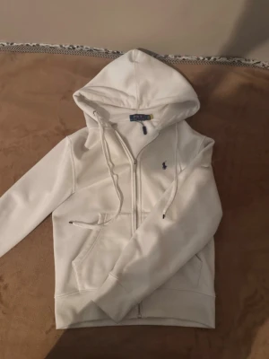 Vit hoodie från Polo Ralph Lauren - Snygg vit hoodie från Polo Ralph Lauren med dragkedja, huva med dragsko och fickor framtill. Klassisk liten logga broderad på bröstet. Mjuk insida i fleece som gör den extra skön att ha på sig. Perfekt för en chill och stilren look.