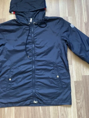 Moncler winbreaker  - Snygg marinblå vindjacka från Moncler med huva och dragsko. Jackan har två framfickor med tryckknappar, hel dragkedja och Moncler-logga på ärmen. Tillverkad i lätt, glansigt material som står emot vind. Perfekt för blåsiga dagar.