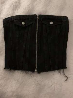 Svart jeanskorsett från Zara - Cool svart korsettopp i jeans från Zara med dragkedja framtill, dekorativa fickor och rå kant nertill. Perfekt att styla med höga jeans eller kjol för en edgy look.