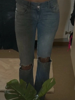Blå jeans med hål vid knäna - Säljer ett par blå jeans med raka ben och stora hål vid båda knäna. Jeansen har klassisk femficksdesign och en ljus tvätt. Bakfickorna har lock och knappdetalj. Perfekta för en avslappnad och trendig look.