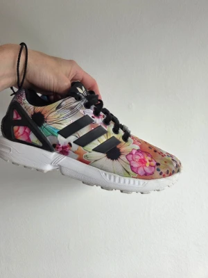 Blommiga sneakers från Adidas - Färgglada sneakers från Adidas med blommigt mönster i rosa, orange, grönt och svart. Skorna har vita sulor, svarta detaljer och klassiska tre ränder på sidan. Ovandelen är i textil och sulan i gummi. Perfekta för dig som vill sticka ut med din stil.