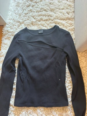 Svart ribbad långärmad topp Gina Tricot - Säljer en svart ribbad långärmad topp från Gina Tricot. Toppen har en cool asymmetrisk detalj över bröstet och är i stretchigt material som sitter snyggt på kroppen. Perfekt att styla med jeans eller kjol för en trendig look.