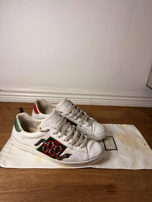 Ace skor  - Vita sneakers från GG med ikonisk broderad röd och svart orm på sidan. Skorna har gröna och röda detaljer på hälen i ormskinns-look och klassisk låg silhuett. Tillverkade i läder med vita snören och gummisula. Storlek 40 passar även 41. Original box finns med två dustbags!