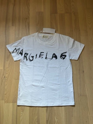 Vit Margiela t-shirt med tryck - Säljer en vit t-shirt från Maison Margiela med svart grafiskt tryck över bröstet där det står 'Margiela 6'. Klassisk passform och rund hals. Tillverkad i mjuk bomull, perfekt till jeans eller shorts.