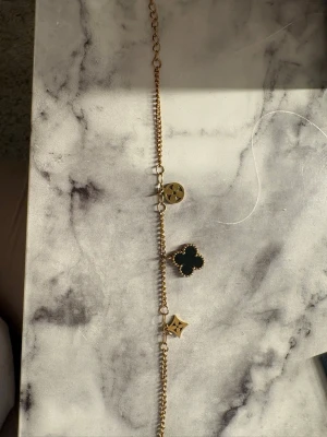 Guldarmband med svarta detaljer - Snyggt guldarmband med tre olika berlocker: en svart fyrklöver, en rund guldberlock och en stjärnformad detalj. Kedjan är tunn och elegant, och armbandet har en justerbar längd. Perfekt accessoar för att lyfta din stil. Fejk