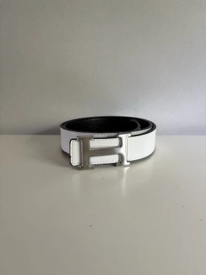 Vitt skärp med silverfärgat H-spänne - Snyggt vitt skärp med ett stort H-format spänne i silverfärg. Skärpet har en stilren design och är gjort i slätt skinn. Insidan är svart vilket ger en cool kontrast. Perfekt accessoar för att lyfta din outfit.