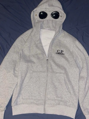 Grå hoodie med glasögon från C.P. Company - Säljer en grå zip hoodie från C.P. Company med ikoniska glasögon i huvan och logga på bröstet. Hoodien har fickor framtill och ribbade muddar. Perfekt för dig som vill sticka ut med en unik detalj i streetwear-stil.