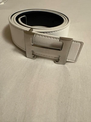 Vitt och svart skärp med H-spänne - Snyggt vändbart bälte i vitt och svart med ett stort H-format metallspänne. Bältet har en stilren design och passar perfekt till jeans eller kostymbyxor. Materialet är slätt och ger en exklusiv känsla.