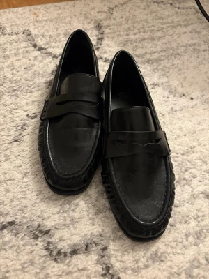 Svarta klassiska loafers i skinn - Säljer ett par stilrena svarta loafers med klassisk penny strap över vristen. Skorna har en tidlös design med rundad tå och låg klack, perfekta för en clean och snygg look. Passar till många olika outfits och är riktigt bekväma att bära. Använda endast 1 gång. 