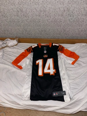 Cincinnati Bengals NFL tröja nr 14 - Säljer en svart, orange och vit Cincinnati Bengals NFL-tröja med nummer 14 och namnet Dalton på ryggen. Tröjan har sportig design med tiger-randiga detaljer på axlarna och är tillverkad i ett lätt och ventilerande material.