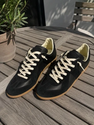 Maison Margiela Gats Svarta Skor Low  - Snygga svarta sneakers med beige sula och vita snören. Ovandelen är i en mix av mocka och slätt läder, vilket ger en stilren och klassisk look. Insidan är ljus och skon har en låg profil som passar till många olika outfits. Ett par feta skor från MM i storlek 44.