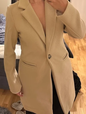 Beige klassisk rock med knäppning - Stilren beige rock med en knapp framtill och två fickor. Rak passform med klassisk krage och helfodrad insida. Perfekt för dig som vill ha en tidlös och enkel look. Materialet ser ut att vara mjukt och lätt strukturerat.