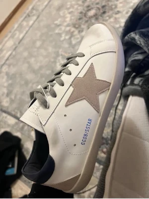Vita sneakers med beige stjärna - Vita sneakers med beige mockastjärna på sidan, grå skosnören och mörkblå detalj vid hälen. Texten GGDB/STAR i blått på sidan. Låg modell med vit sula och stilren design som passar till streetwear och casual outfits.