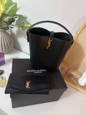 Svart bucket bag  - Säljer en svart bucket bag i slätt läder med guldfärgad YSL-logga framtill. Väskan har ett stilrent handtag och klassisk form. Perfekt för dig som gillar lyxiga accessoarer med elegant design.