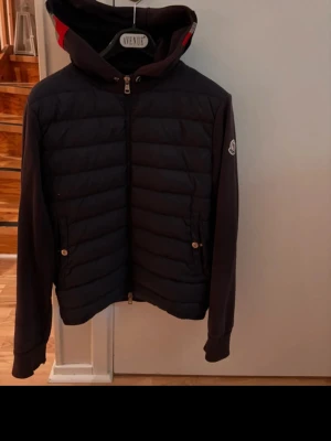 Moncler cardigan - Svart cardiganjacka från Moncler med vadderad framsida och mjuka ärmar. Jackan har huva med röd och grå rand, dragkedja framtill och två fickor med knapp. Moncler-logga på ärmen ger en exklusiv touch. Perfekt för kyliga dagar.