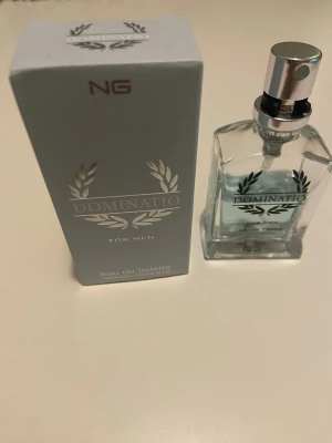 Dominatio Eau de Toilette för män - Dominatio Eau de Toilette för män med fräsch och maskulin doft. Flaskan är genomskinlig med silverdetaljer och har en stilren, rektangulär form. Förpackningen är grå med silvertext och dekorativa lagerblad.