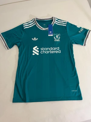 Turkos Liverpool matchtröja Adidas - Säljer en turkos Liverpool matchtröja från Adidas med vita detaljer och klubbmärke på bröstet. Tröjan har korta ärmar, rundad krage med svart och vit rand samt Adidas klassiska ränder på axlarna. Tillverkad i lätt funktionsmaterial.