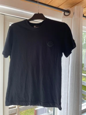 Svart Moncler t-shirt med logga - Snygg svart t-shirt från Moncler med diskret svart logga på bröstet. Klassisk rund halsringning och korta ärmar. Tillverkad i mjuk bomull som känns skön mot huden. Perfekt till jeans eller shorts för en clean look.