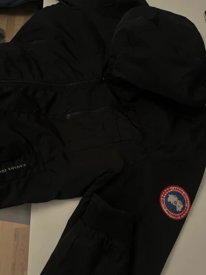 Svart vindjacka från Canada Goose - Säljer en svart vindjacka från Canada Goose med huva och dragkedja. Jackan har en tydlig logotyp på ärmen och praktiska fickor. Materialet är tunt och vindtätt, perfekt för blåsiga dagar. Snygg och funktionell design som passar till det mesta.