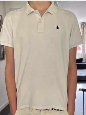 Vit pikétröja från Morris Stockholm - Klassisk vit pikétröja från Morris Stockholm med korta ärmar och liten broderad logga på bröstet. Tröjan har krage och knappar framtill. Perfekt till jeans eller shorts för en clean look. (Säljer eftersom den är för liten)