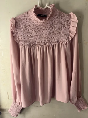 Ljusrosa blus - Supersöt ljusrosa blus med smock upptill, volang vid axlar och hals samt långa puffiga ärmar med smockade muddar. Tillverkad i ett tunt, flowy tyg som ger en romantisk vibe. Perfekt för dig som gillar feminina detaljer och en trendig look. 