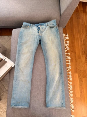 Levi's 501 ljusblå raka jeans - Klassiska Levi's 501 jeans i ljusblå tvätt med raka ben och knappgylf. Jeansen har fem fickor, tydliga sömmar och den ikoniska läderpatchen bak i midjan. Perfekta för en avslappnad och tidlös stil.