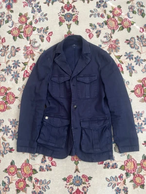 Hansen & Jacob safarijacka - Snygg mörkblå kavaj i utility-stil med fyra stora fickor framtill och klassisk krage. Rak passform och knappstängning framtill. Perfekt för dig som gillar en avslappnad men stilren look. Storlek M nypris 4000