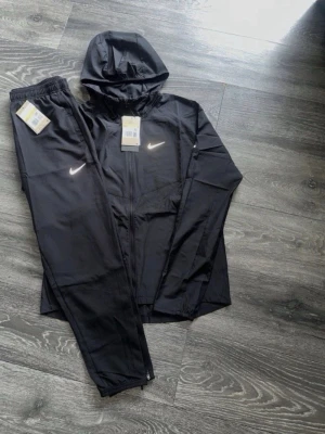 Svart träningsset från Nike med huva - Snyggt svart träningsset från Nike med jacka och byxor. Jackan har huva, dragkedja och två fickor samt Nike-logga på bröstet. Byxorna har resår i midjan och Nike-logga på benet. Perfekt för träning eller sportiga dagar.