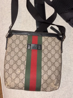 Gucci axelväska med GG-mönster - Säljer en klassisk Gucci axelväska i beige canvas med GG-monogram, grönt och rött band i mitten och svarta läderdetaljer. Väskan har justerbar axelrem och präglad logotyp på framsidan. Perfekt för dig som gillar ikoniska designeraccessoarer.