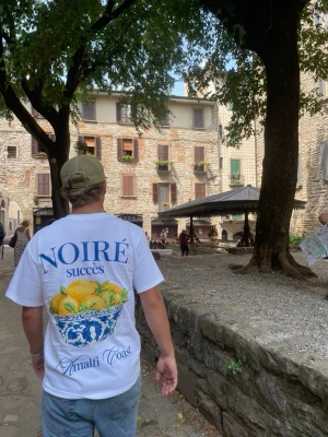 Noiré T-shirt - Säljer en vit t-shirt med stort färgglatt tryck på ryggen med citroner i en blåvit skål och texten 'NOIRÉ succès Amalfi Coast'. T-shirten har korta ärmar och en relaxed passform. Perfekt för dig som gillar statement-plagg och somriga vibes. Finns alla storlekar och fraktar i hela Sverige. Noiresverige.se finns vi också på!