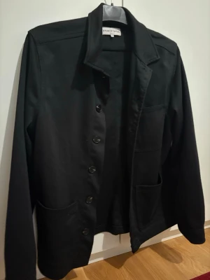 Svart overshirt från Studio Total - Stilren svart overshirt från Studio Total med klassisk krage, knäppning framtill och tre praktiska fickor. Overshirten har raka ärmar och en avslappnad passform, perfekt att bära över en t-shirt eller hoodie. Materialet ser ut att vara bomull eller bomullsblandning.