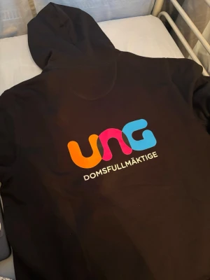 Svart hoodie UNG Domstolsfullmäktige - Svart hoodie från South West i storlek XL med färgglad UNG Domstolsfullmäktige-tryck på ryggen. Klassisk huva och mjukt material, perfekt för dig som vill sticka ut med en unik och bekväm stil. Materialet är bomullsmix.