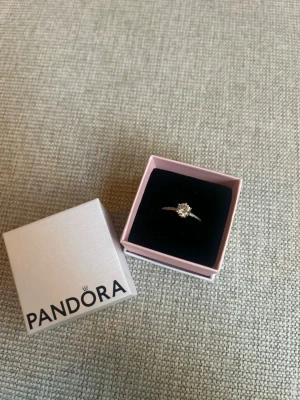 Guldfärgad ring med sten från Pandora - Snygg guldfärgad ring från Pandora med en stor klar sten i mitten och små gnistrande stenar längs bandet. Ringen har en elegant och klassisk design som verkligen sticker ut. Perfekt för dig som vill ha något extra till din stil.