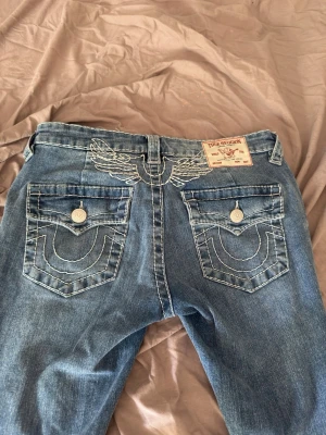 Blå True Religion jeans med brodyr - Säljer ett par blå jeans från True Religion med ikonisk vit brodyr på bakfickorna och vingdetalj vid linningen. Klassisk femficksmodell med raka ben och tydliga kontrastsömmar. Perfekta för dig som gillar streetwear och vill sticka ut. Nästintill aldrig använda så väldigt bra skick, köptes för 1800 därav priset💞