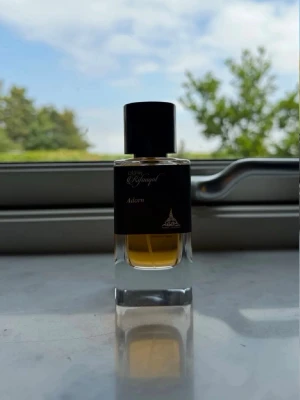 Svart parfymflaska Adorn - Säljer en stilren parfymflaska med svart lock och etikett. Flaskan är rektangulär i genomskinligt glas och innehåller en gyllene vätska. Märkt med 'Adorn' och elegant designad för dig som gillar exklusiva dofter.