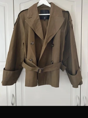 Olivgrön kort trenchcoat med bälte - Stilren olivgrön trenchcoat med dubbelknäppning, breda slag och axelklaffar. Jackan har ett matchande bälte i midjan och justerbara ärmslut. Kortare modell som ger en modern look. Perfekt för lager-på-lager och snygg till jeans eller kjol.