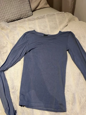 Blå långärmad topp - En enkel och stilren blå långärmad topp med rund hals. Toppen har en smal passform och är gjord i ett mjukt, tunt material som känns skönt mot huden. Perfekt att matcha med jeans eller kjol för en clean look.