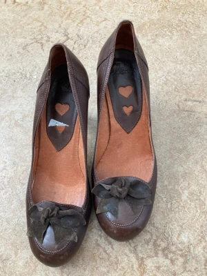 Bruna pumps med rosett och hjärtdetalj - Snygga bruna pumps med rund tå och dekorativa rosetter framtill. Skorna har unika hjärtformade detaljer på innersulan och är tillverkade i ett material som ser ut som läder. Perfekta för dig som vill ha en söt och stilren look.