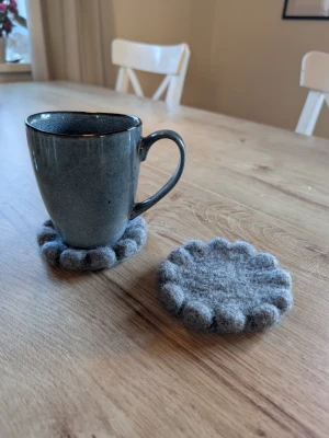 Två stycken gråa glasunderlägg i 100% ull - Två runda glasunderlägg tovade i 100% ull. Handgjorda av mig och är helt nya och oanvända! Ca 11cm i diameter. OBS: muggen ingår ej! 